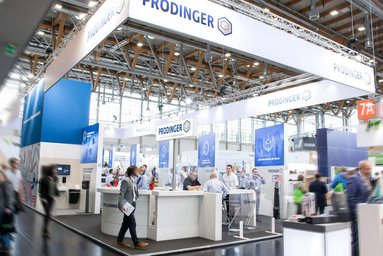 In einer Messehalle ist ein Stand von PRODINGER zu sehen, auf dem Besucher von Mitarbeitern beraten werden.