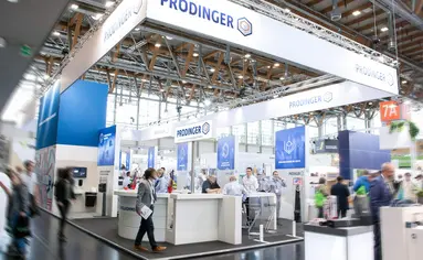 In einer Messehalle ist ein Stand von PRODINGER zu sehen, auf dem Besucher von Mitarbeitern beraten werden.